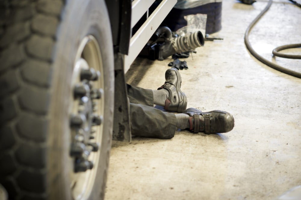 Bildet viser beina og føttene til en mekaniker som ligger under et kjøretøy for å utføre reparasjoner. Personen har på seg slitte arbeidsklær og sikkerhetssko, og man kan se verktøy og en slange spredt rundt på det oljeflekkete verkstedgulvet.